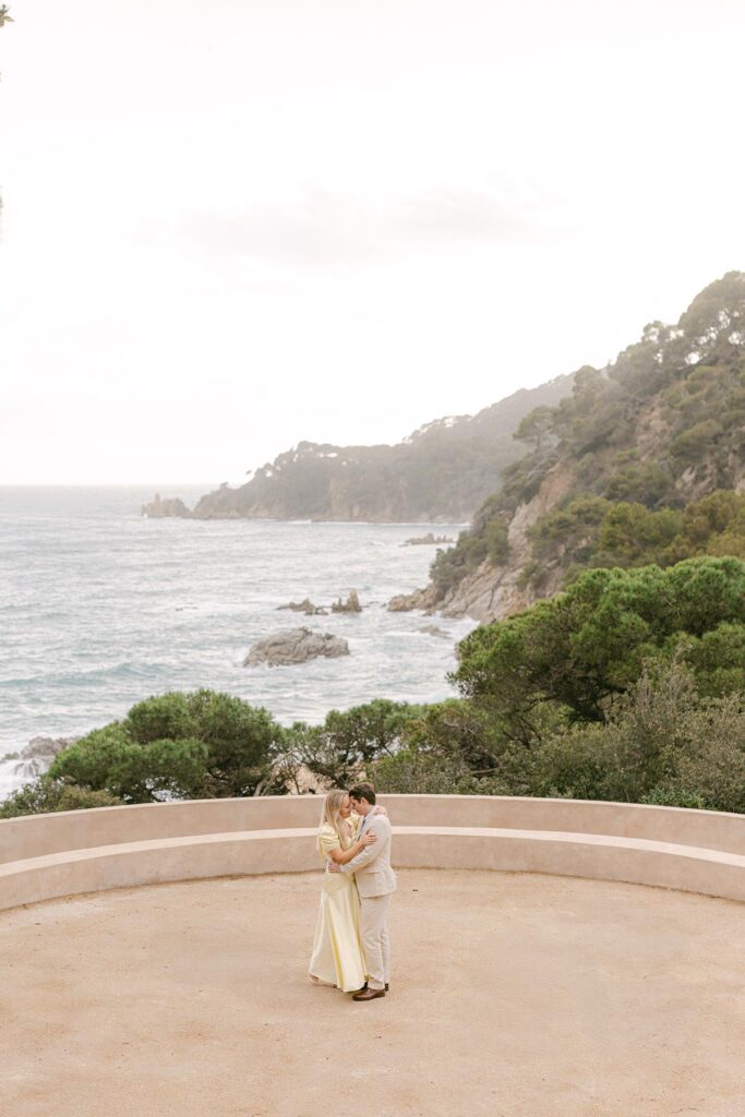 Couple embracing on a circular stone terrace overlooking the Mediterranean Sea at Santa Clotilde Gardens engagement session near Barcelona. Captured by an experienced Barcelona engagement photographer