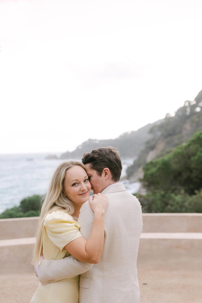 Bride-to-be smiling and embracing her fiancé during a romantic engagement session by the sea in Lloret de Mar