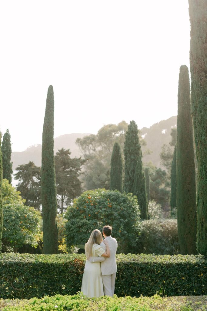 Engaged couple strolling through tree-lined garden path in soft, neutral outfits during romantic photoshoot in Costa Brava, Spain