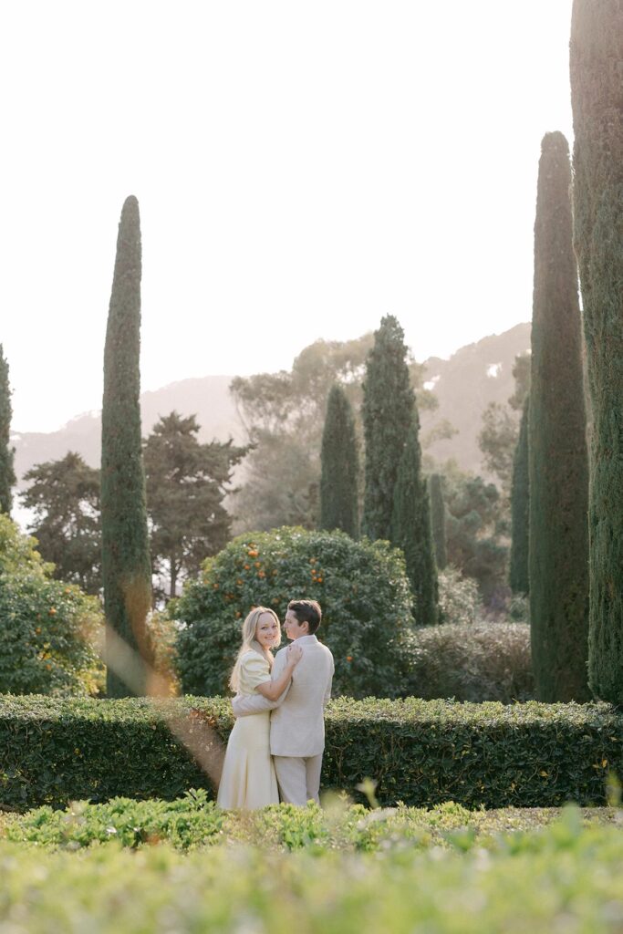 Engaged couple strolling through tree-lined garden path in soft, neutral outfits during romantic photoshoot in Costa Brava, Spain. Captured by an experienced Barcelona engagement photographer