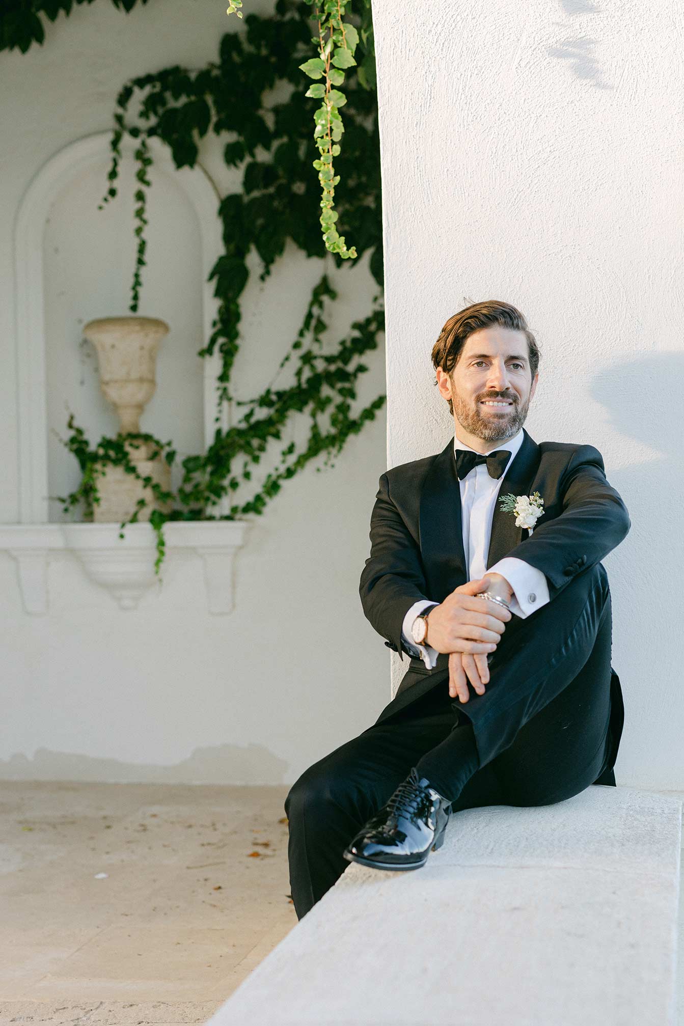 Groom in a black tuxedo sitting on a stone ledge at Hostal La Gavina with Mediterranean architecture in the background